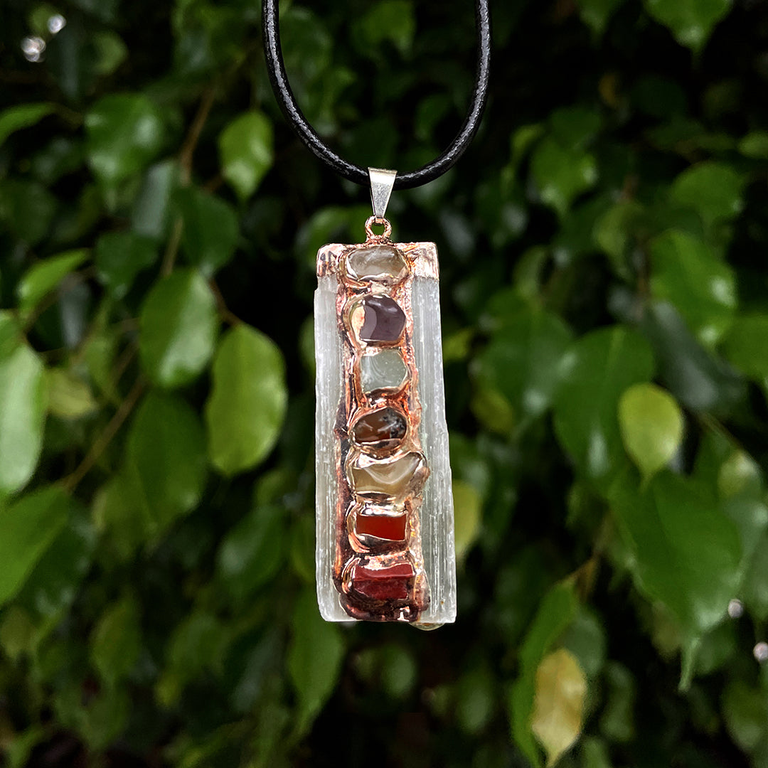 Handcrafted selenite pendant with polished gemstones set in a copper frame, hanging on a black cord against a green leafy background.